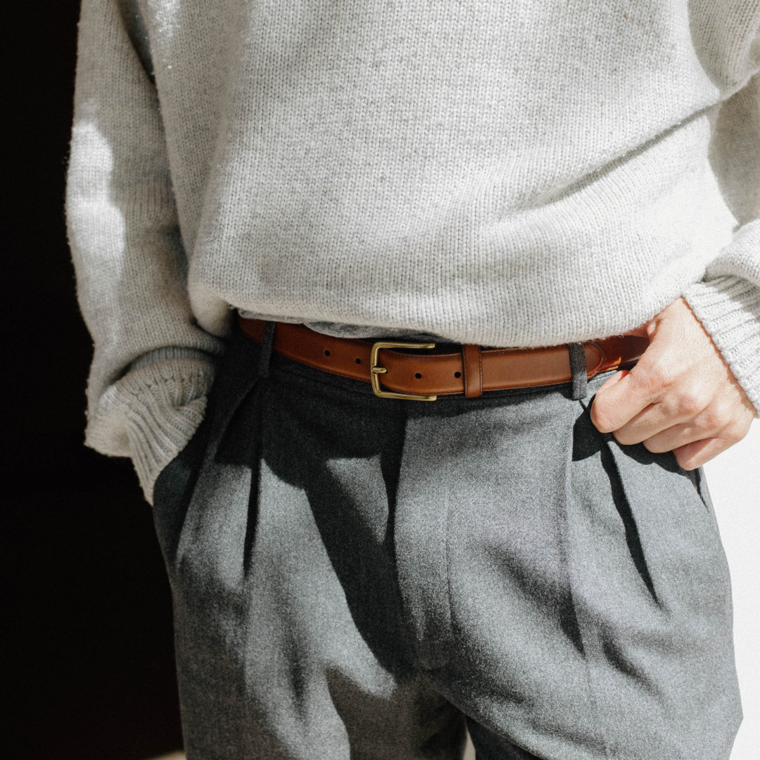 Cognac brown leather belt with brass buckle worn at the waist over gray pleated trousers and cream knit sweater