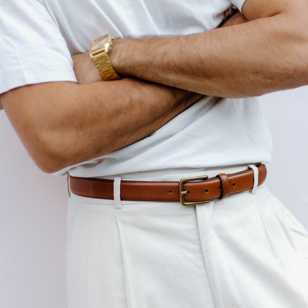 Close-up of a person's crossed arms wearing a gold watch, white shirt, white pants, and a brown leather belt with a brass buckle.