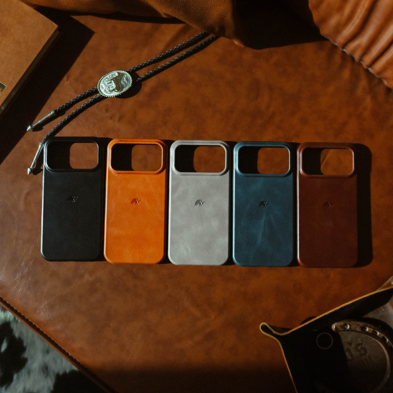 Five leather phone cases (black, orange, gray, teal, brown) side-by-side on brown leather, each bearing a Bullstrap logo.