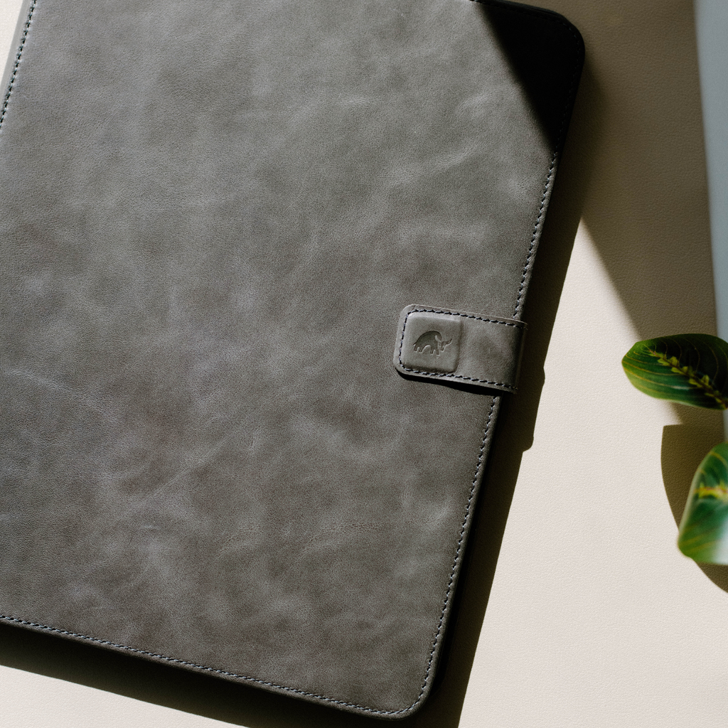 Gray leather iPad case with a clasp, placed on a light surface next to a plant.