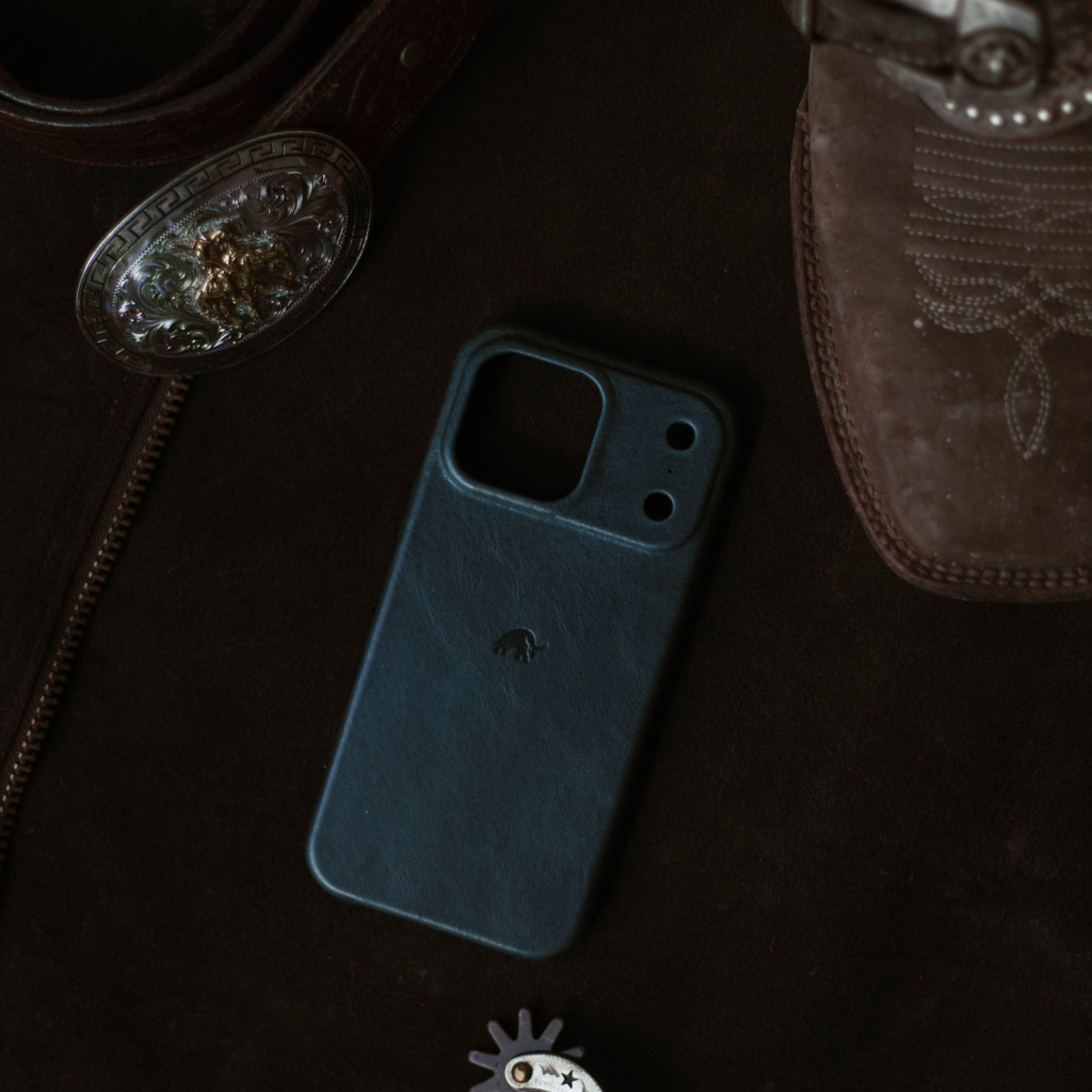 Ocean-blue leather iPhone case with dual camera cutouts lying on dark fabric beside a western belt buckle and leather boot.