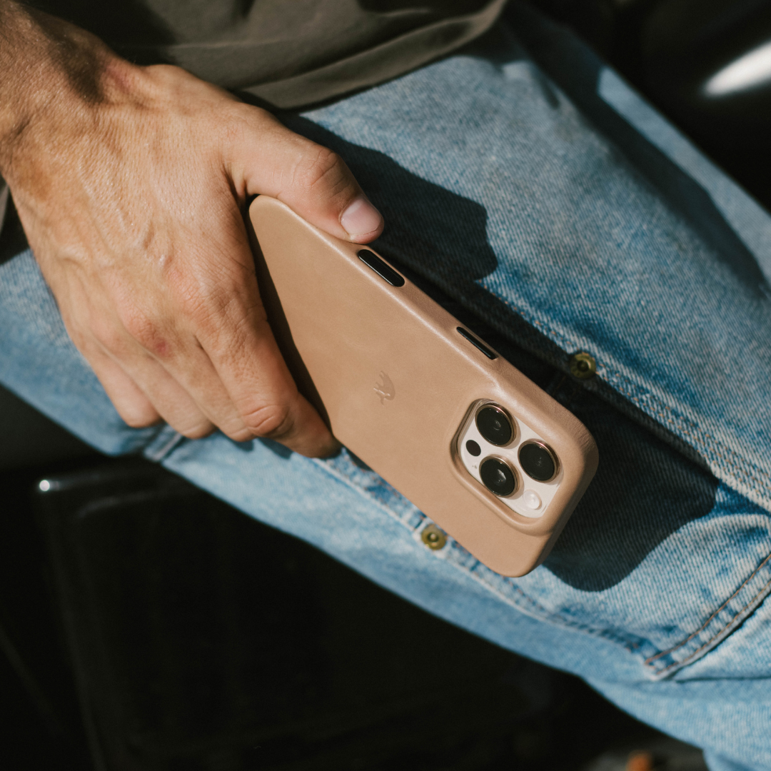 Hand holding phone with tan leather 'Dune' minimalist case, camera cutouts visible, resting on blue jeans