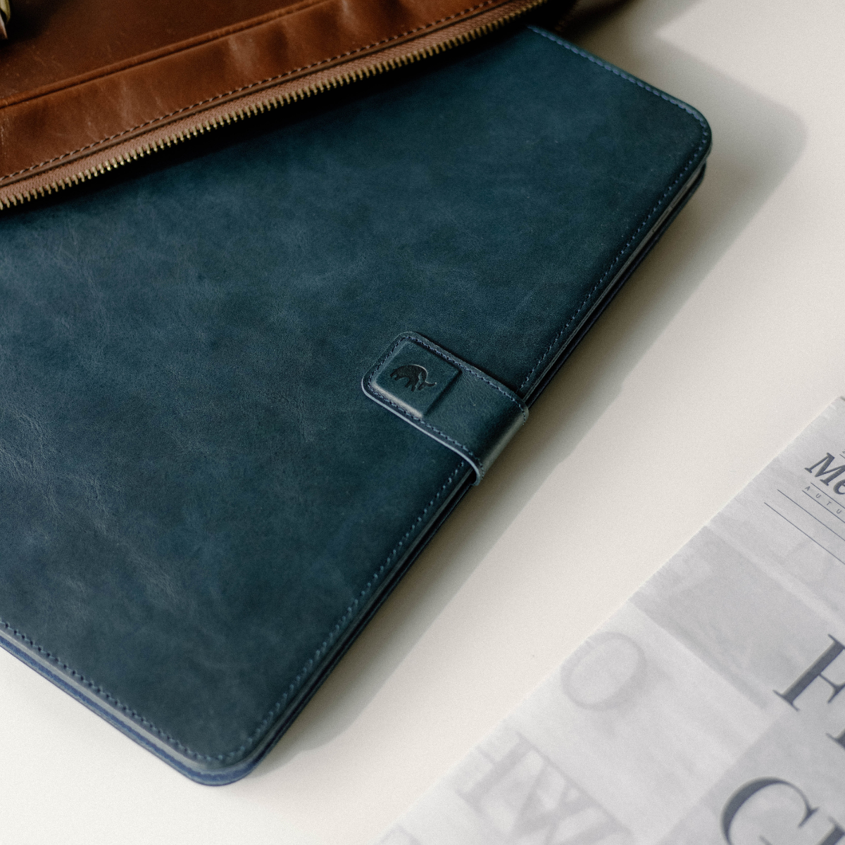 Blue leather tablet folio with strap and embossed elephant logo next to a brown leather zip pouch and folded newspaper.