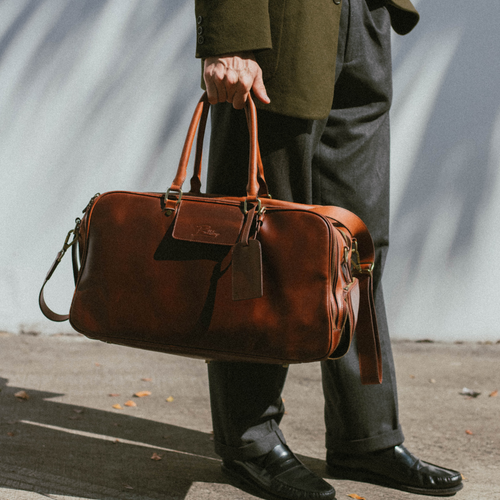 Brown leather weekender bag with top handles and luggage tag, held by a person wearing dark trousers and black loafers.
