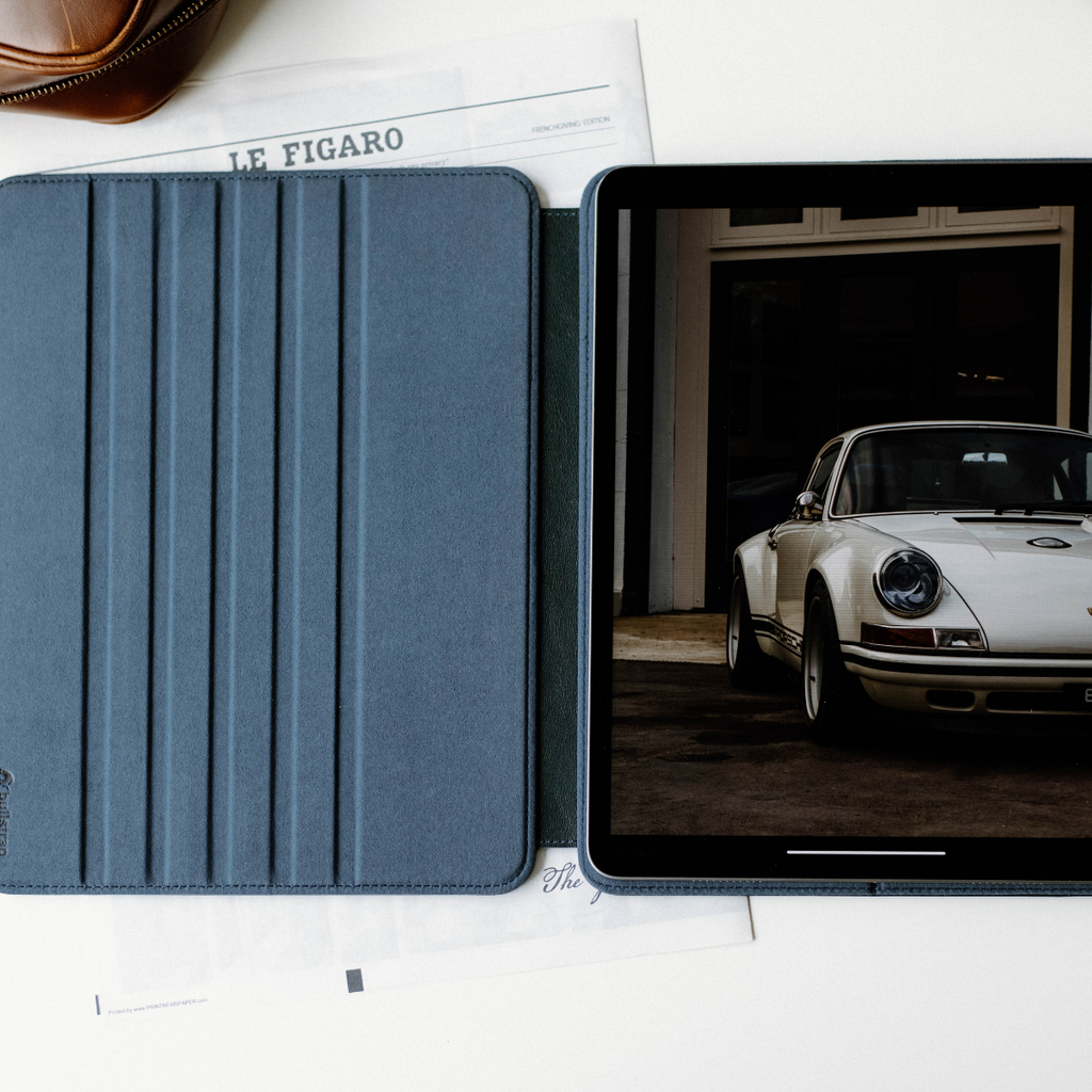 Blue leather iPad case open beside tablet showing car photo; newspaper headline 'LE FIGARO' and partial word 'The' visible