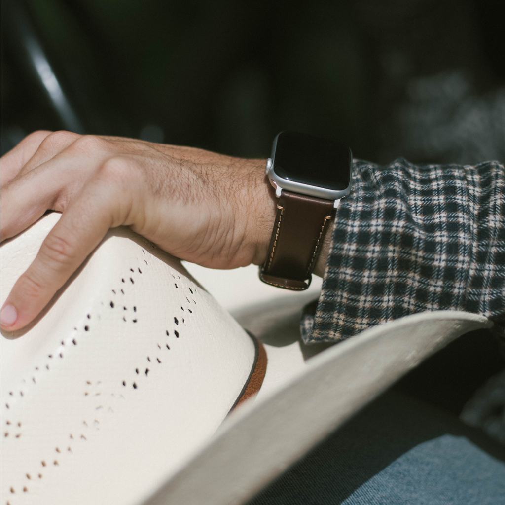 Close-up of wrist wearing Modern Terra dark brown leather Apple Watch strap showing contrast stitching and silver hardware while holding a white hat