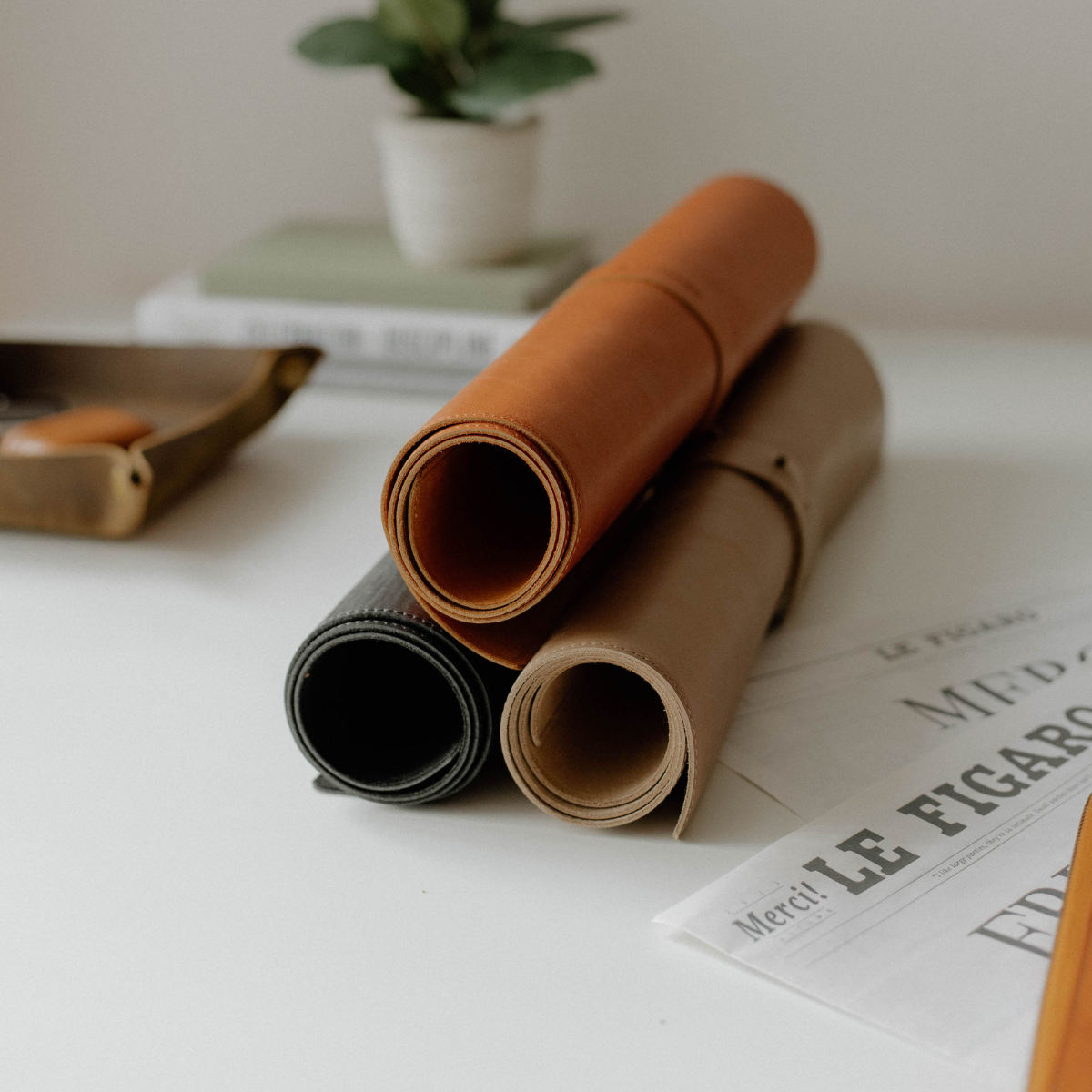 Three rolled leather desk mats in sienna, black, and tan colors on a white table with a plant in the background.