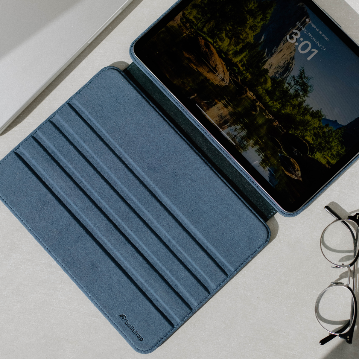 Tablet in blue folio on table showing lock screen '3:01', 'Monday, November 27', 'Swipe up to unlock'; eyeglasses beside.