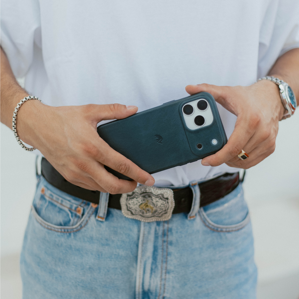 Hands holding an iPhone in a dark teal leather minimalist case; person wearing jeans and a western belt buckle.