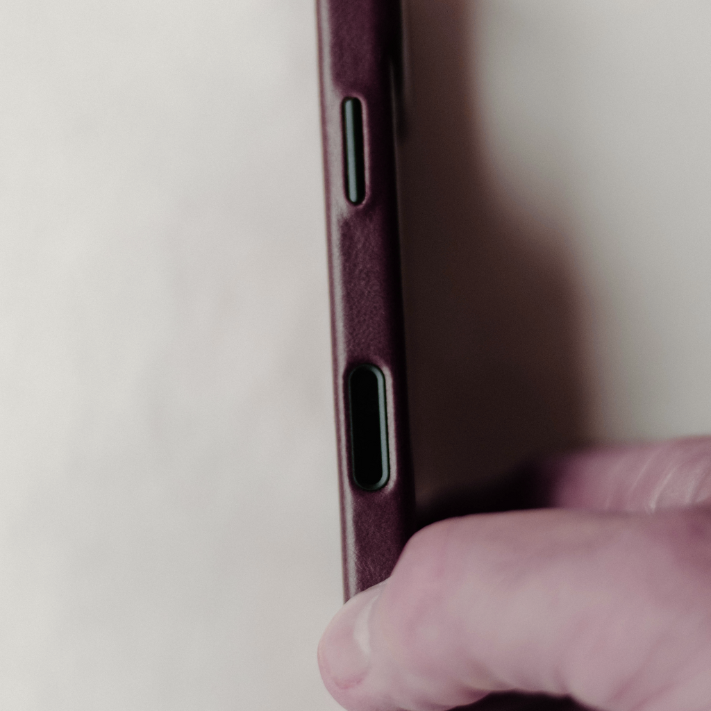 Side edge of bourbon leather phone case showing volume button and speaker cutouts with a thumb holding the bottom edge.