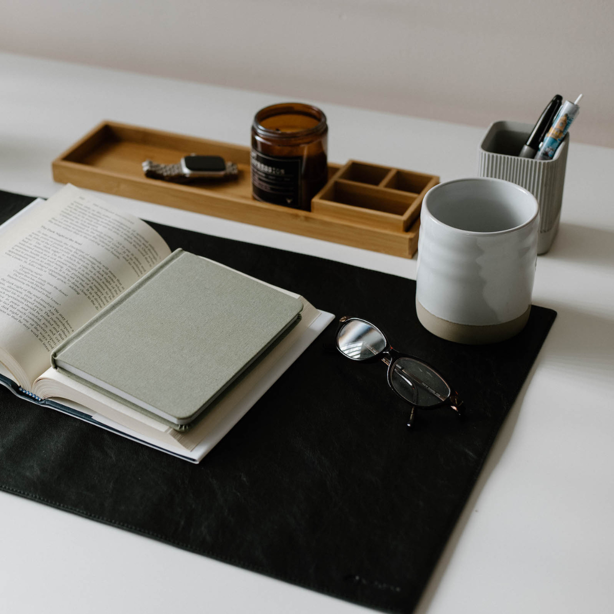 Open book and gray notebook on a black desk mat, reading glasses, ceramic mug, wooden tray with candle and watch, and a pen holder.