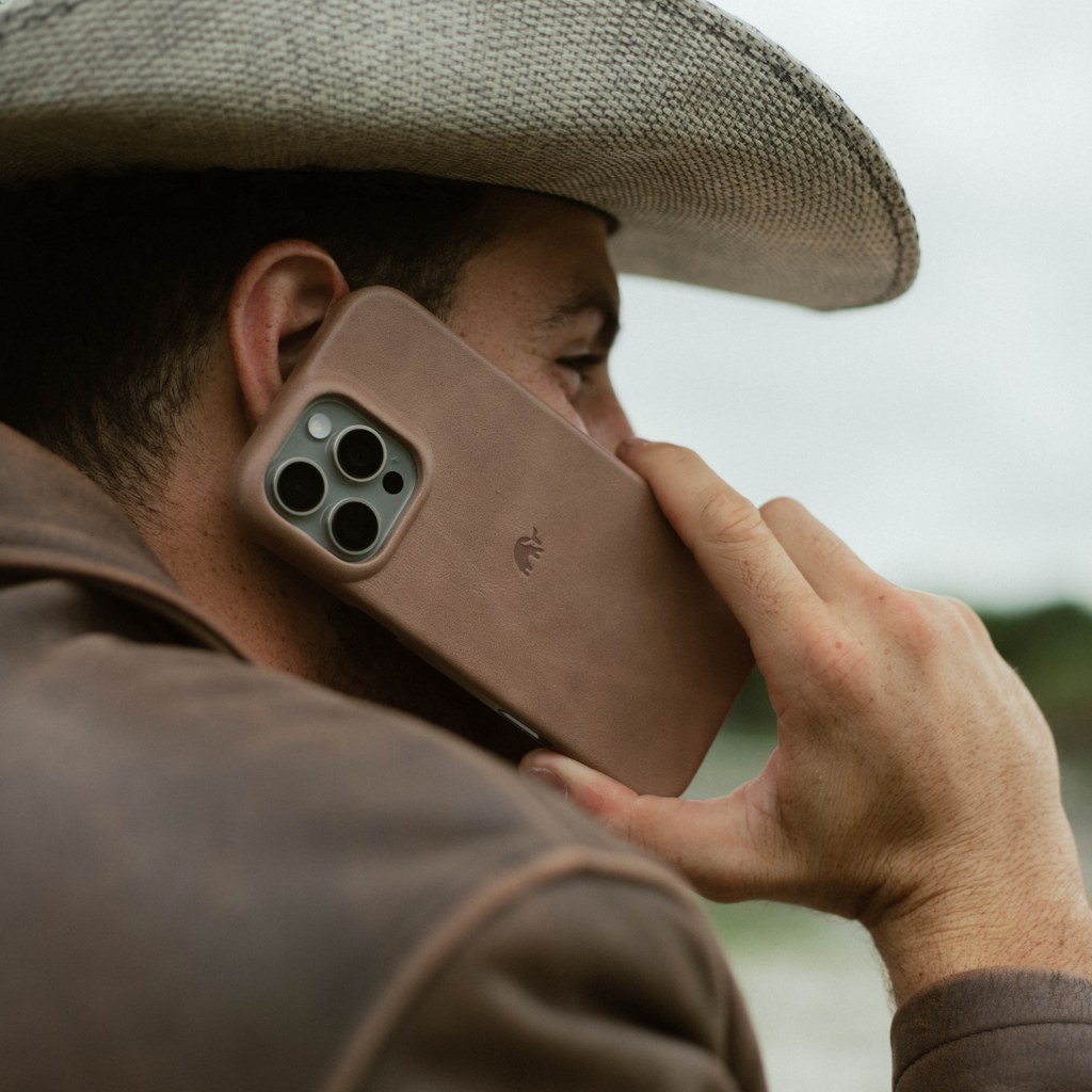 Person in a cowboy hat holding a phone to their ear with a brown leather case showing triple camera and embossed logo
