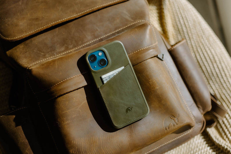 Green leather phone case with card pocket showing 'ONE' on a dollar bill, resting on a brown leather backpack.