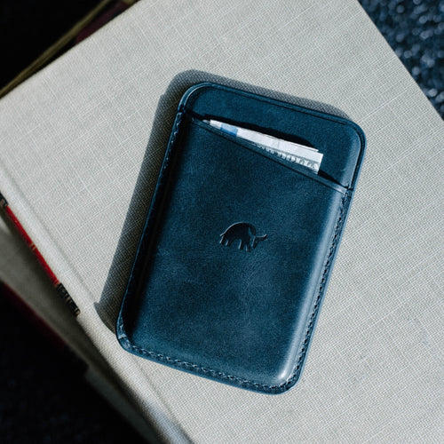 Slim dark-blue leather card wallet with embossed rhino logo and folded cash in top pocket, on a beige textured book.
