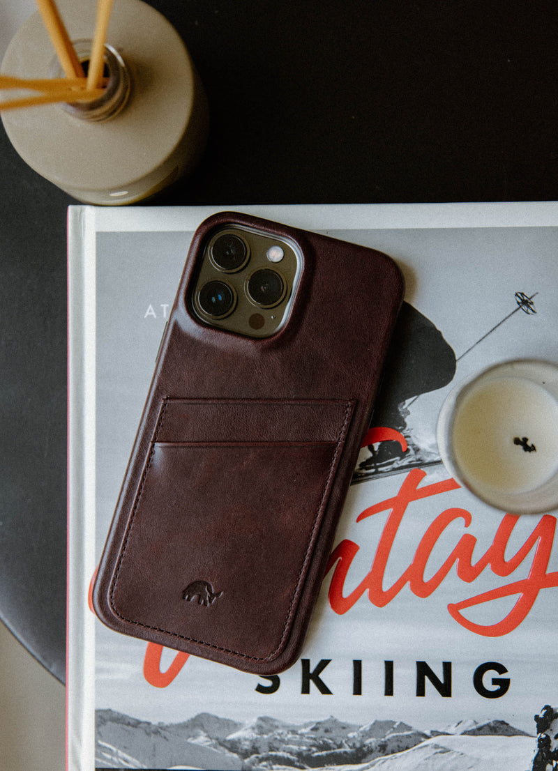 Leather bourbon iPhone case on a book with cover text 'vintage' and 'SKIING', candle and vase in the background.