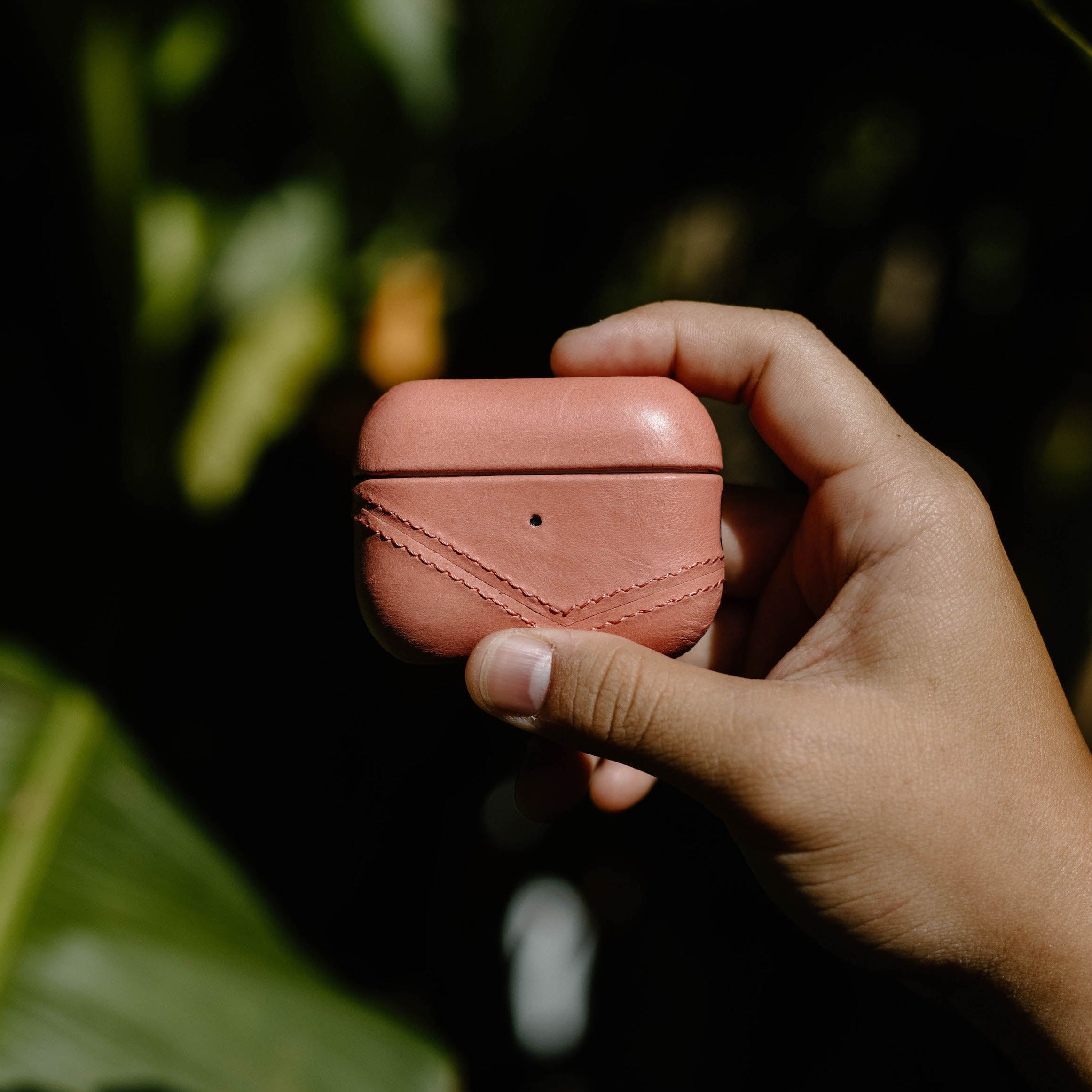 Pink leather AirPods case with diagonal stitching held in a hand against blurred green foliage