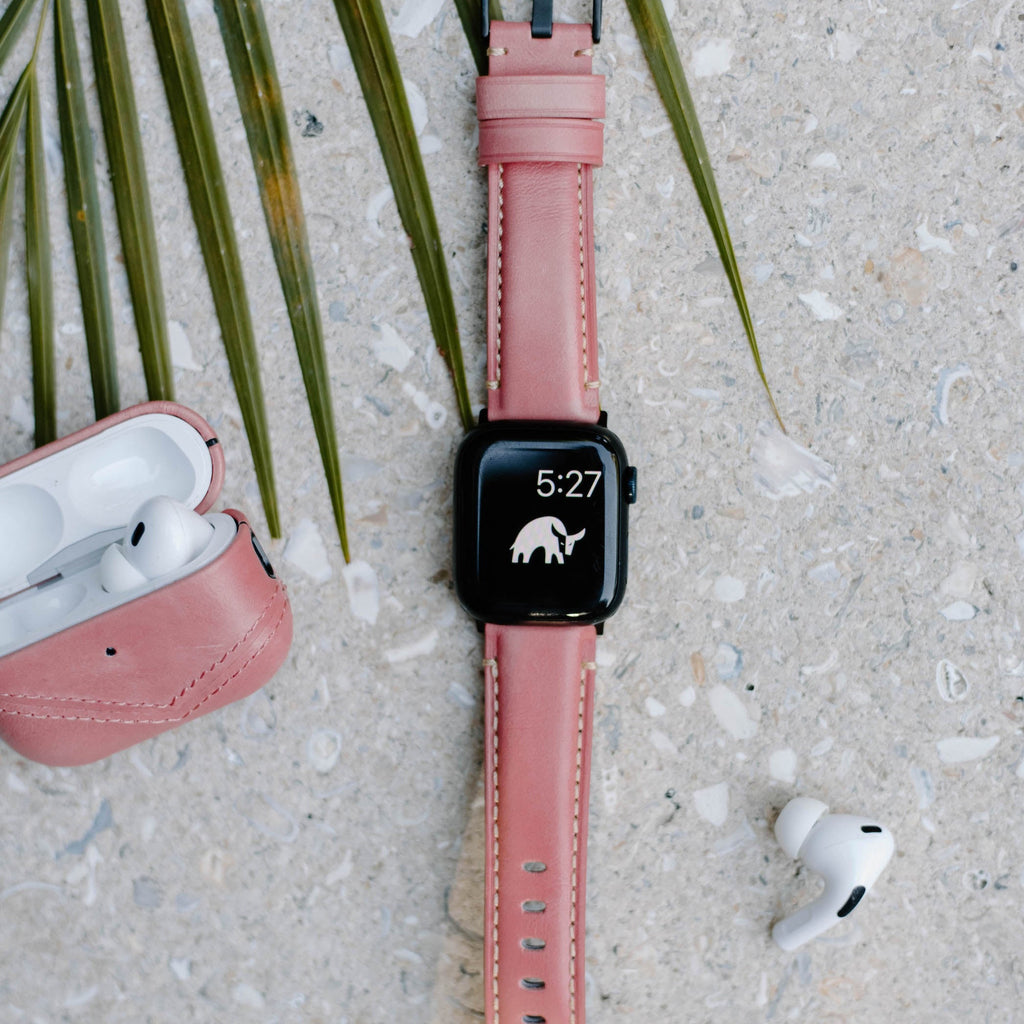 Pink 'South Beach' leather Apple Watch strap on watch displaying 5:27 and elephant icon, beside pink AirPods case and white earbud