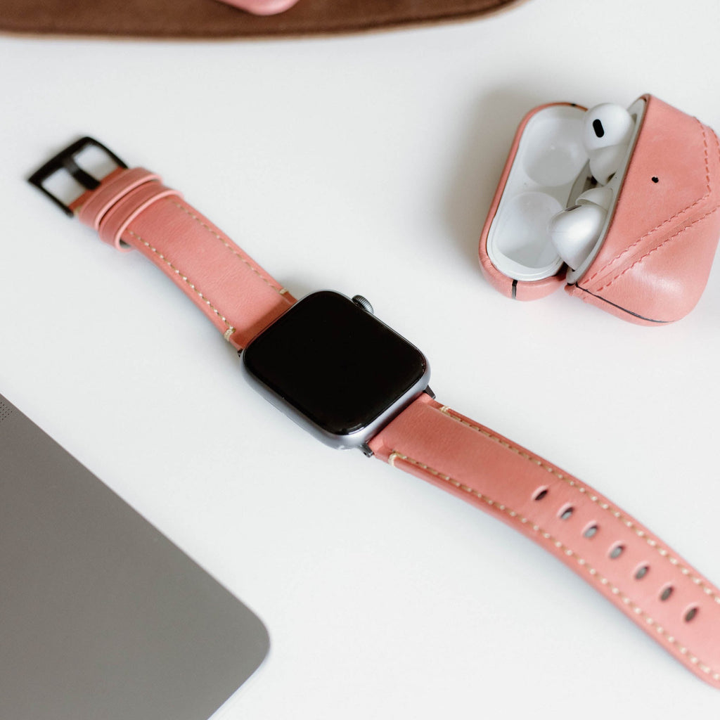 Smartwatch with coral-pink leather band beside open pink earbuds case and laptop corner on white desk.