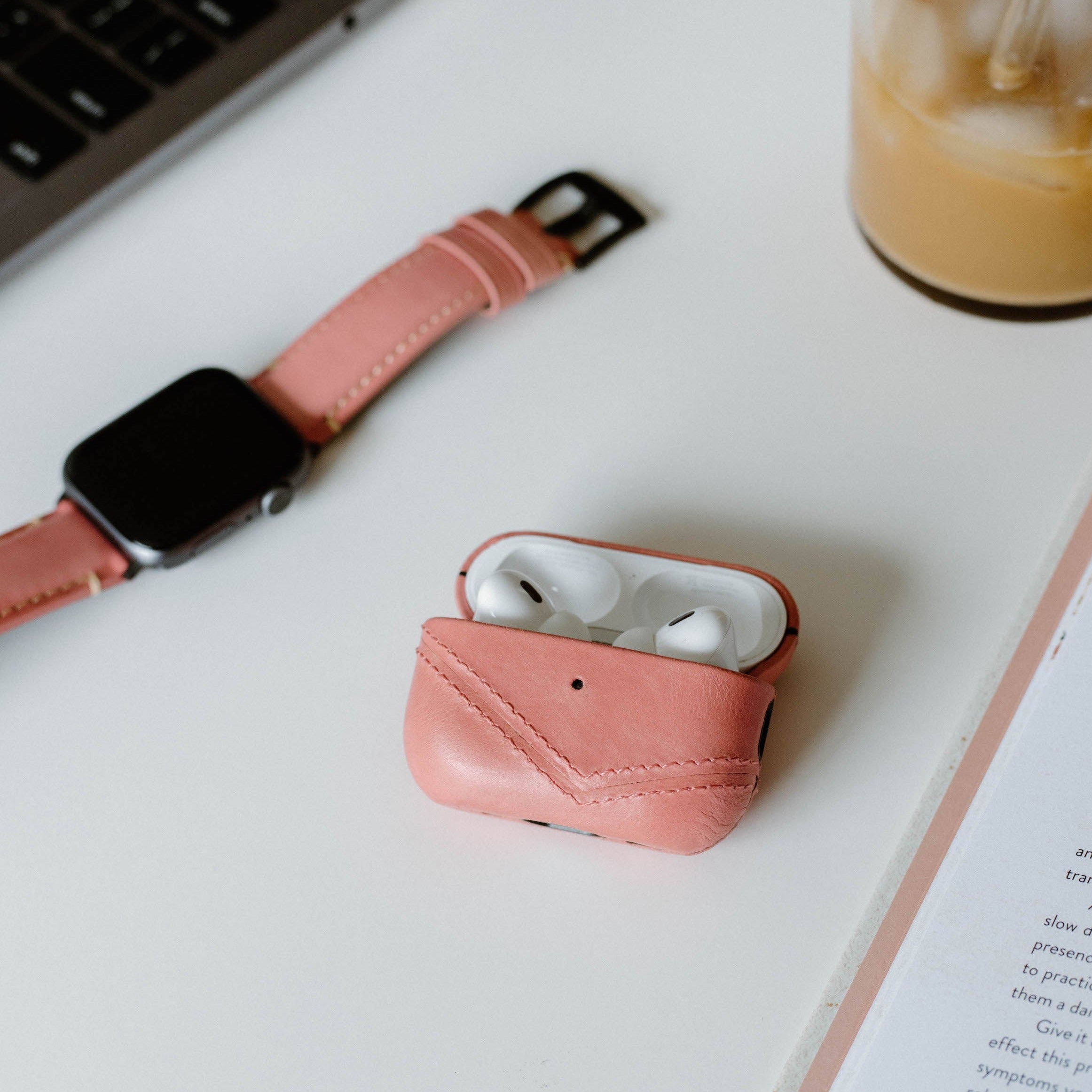 Pink leather AirPods Pro case (SOUTH BEACH) open with earbuds on white desk beside pink watch and iced coffee.