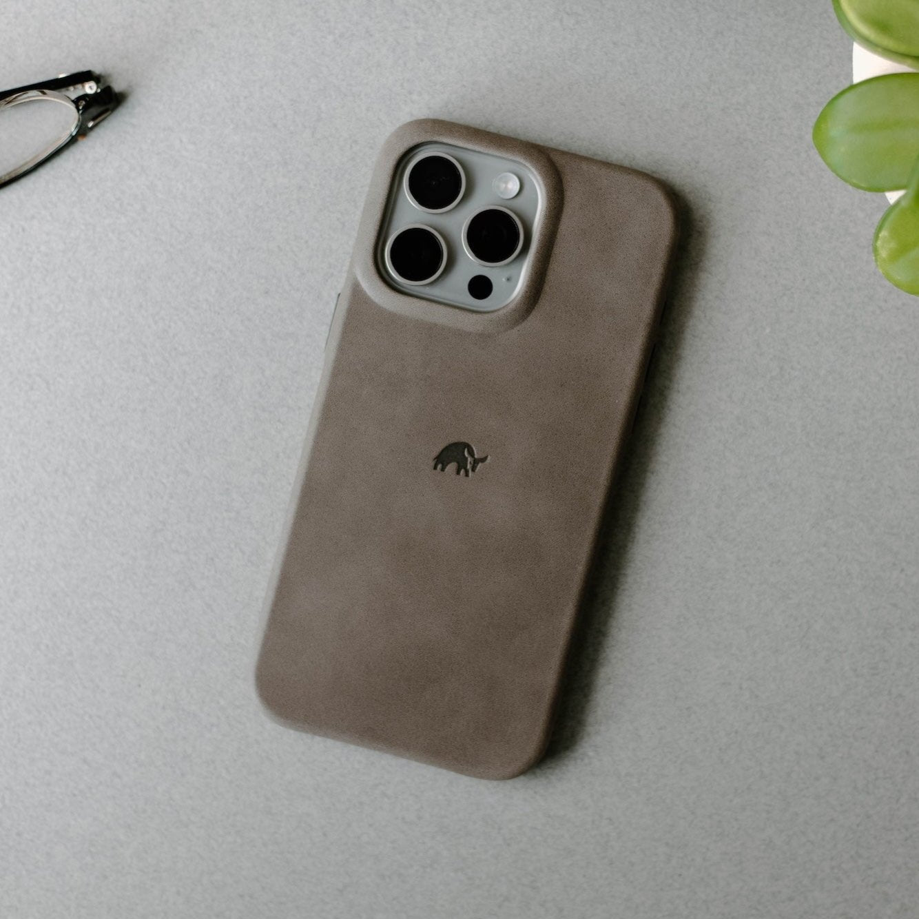 Brown leather iPhone case with triple camera cutout and small elephant logo resting on gray tabletop.
