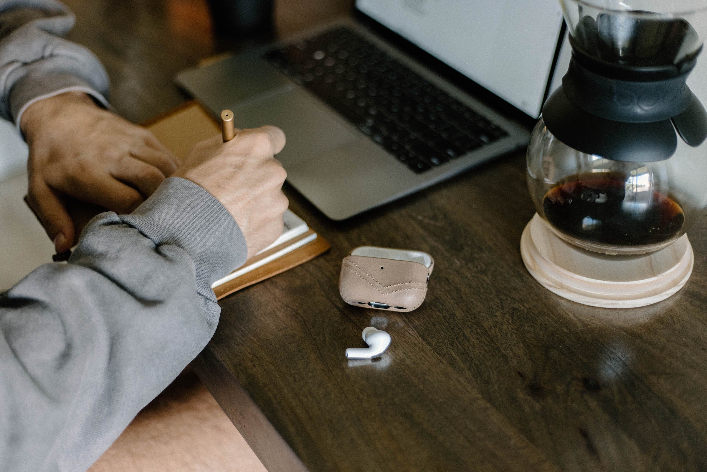 Person writing in a notebook at a wooden table with a laptop, wireless earbud with case, and a glass coffee pot.