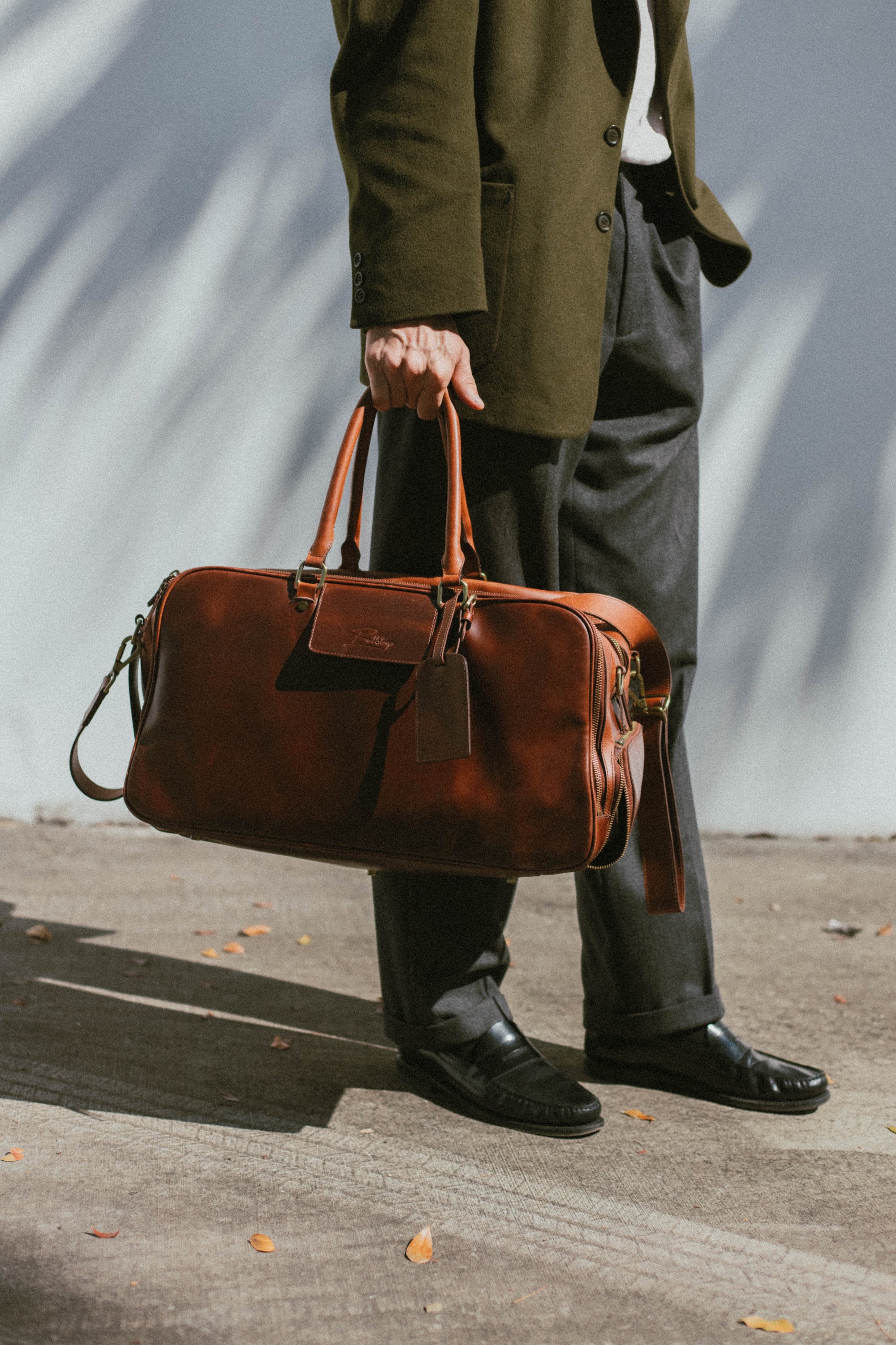 Person holding brown leather weekender bag by the handles, wearing olive coat, gray trousers and black loafers on sunlit pavement.