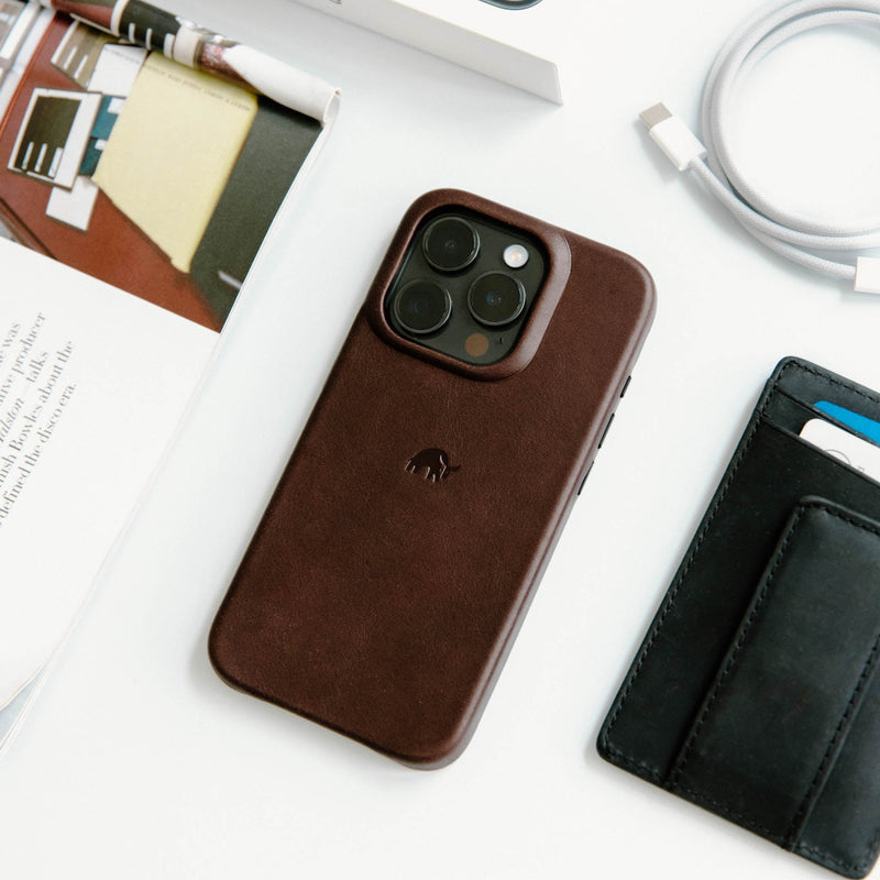 Brown leather iPhone case (triple camera) on white desk with USB‑C cable, magazine pages and black card wallet.