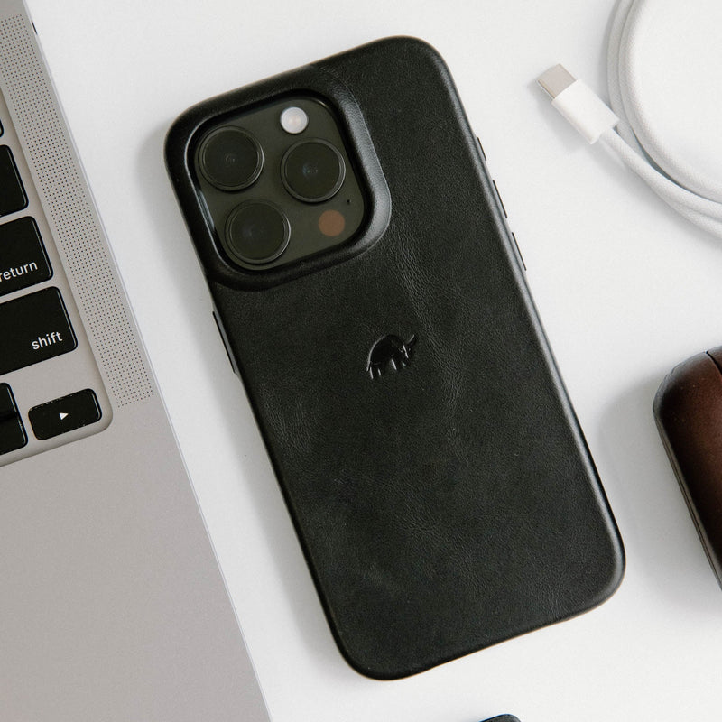 Black Minimalist leather phone case on a white surface beside a laptop, showing keys 'shift' and 'return' and an embossed elephant logo.