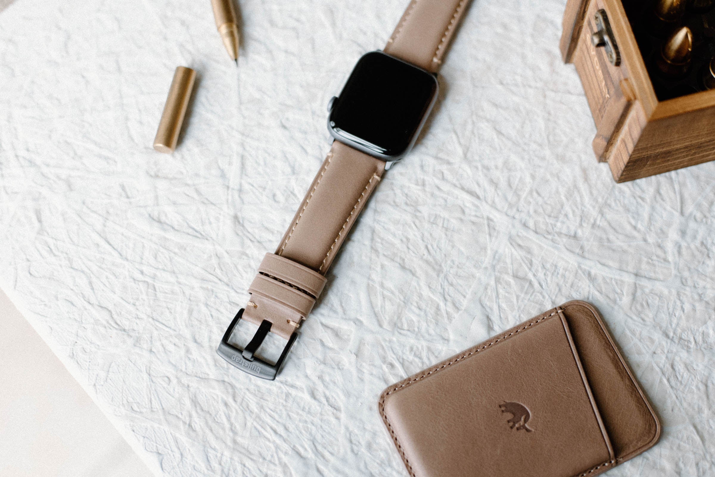 Leather Apple Watch strap in Dune color displayed on a textured surface with a wallet and pen nearby.
