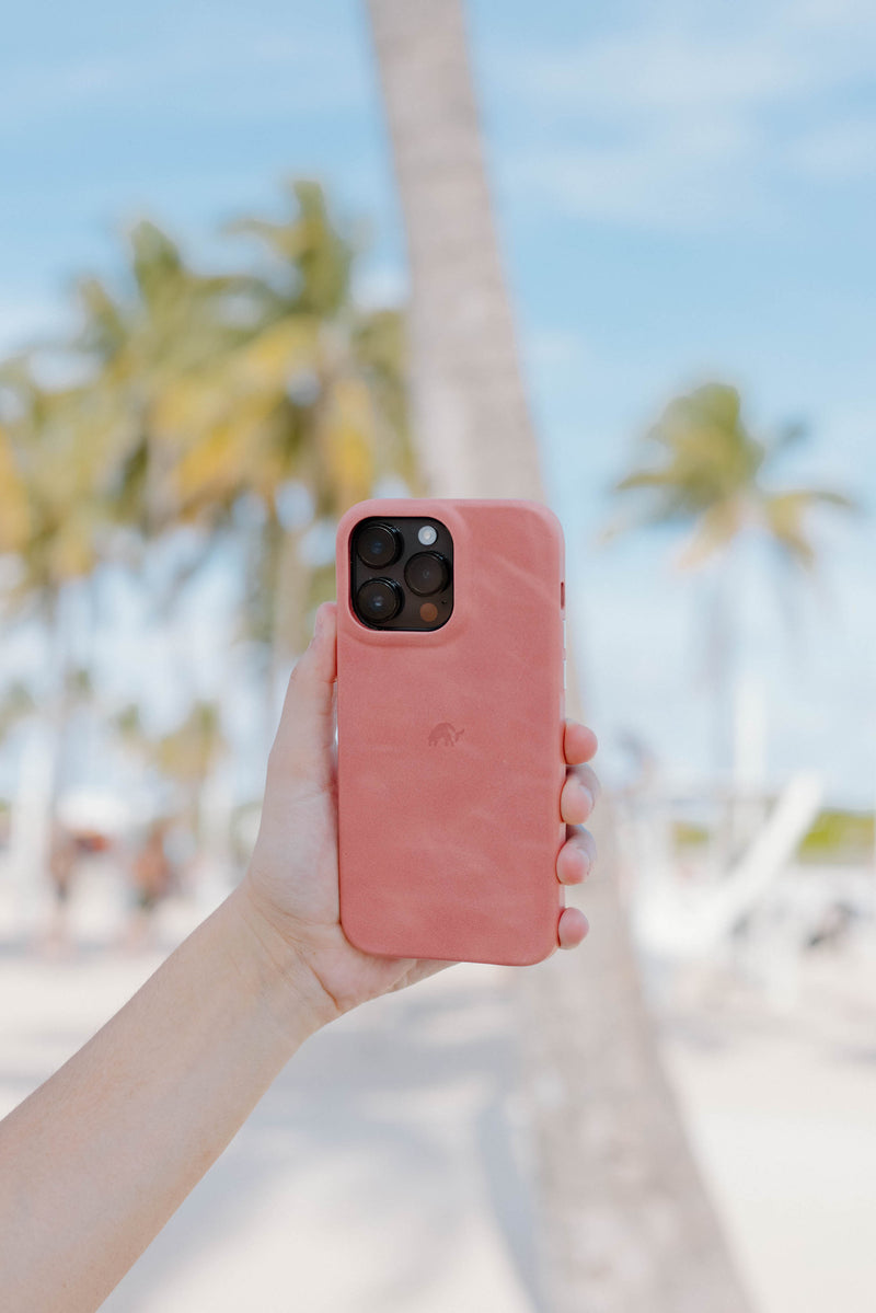 A hand holding a pink Bullstrap iPhone case in a tropical setting with palm trees in the background.