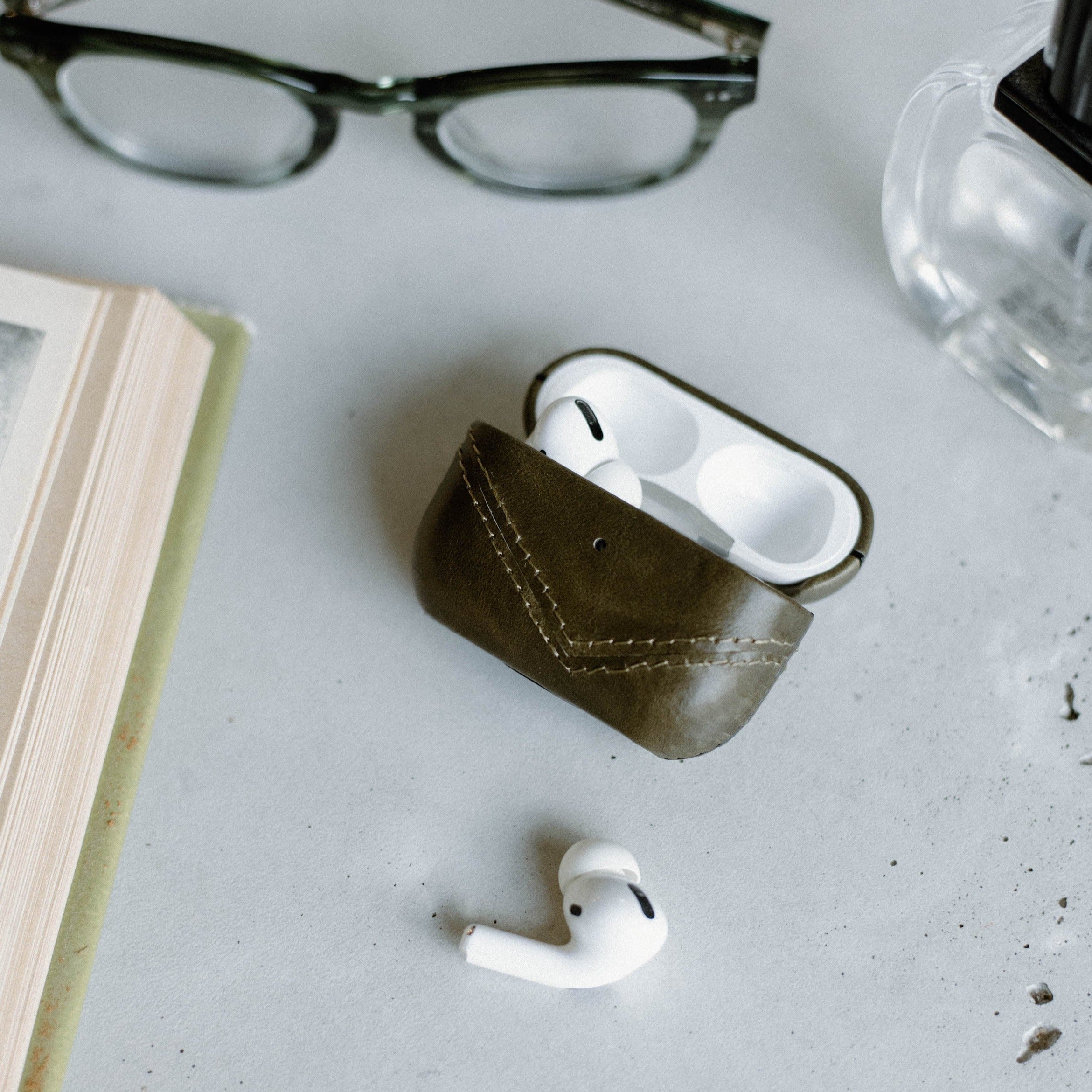 A leather AirPods case in green with AirPods partially visible, placed on a table with a book and glasses.