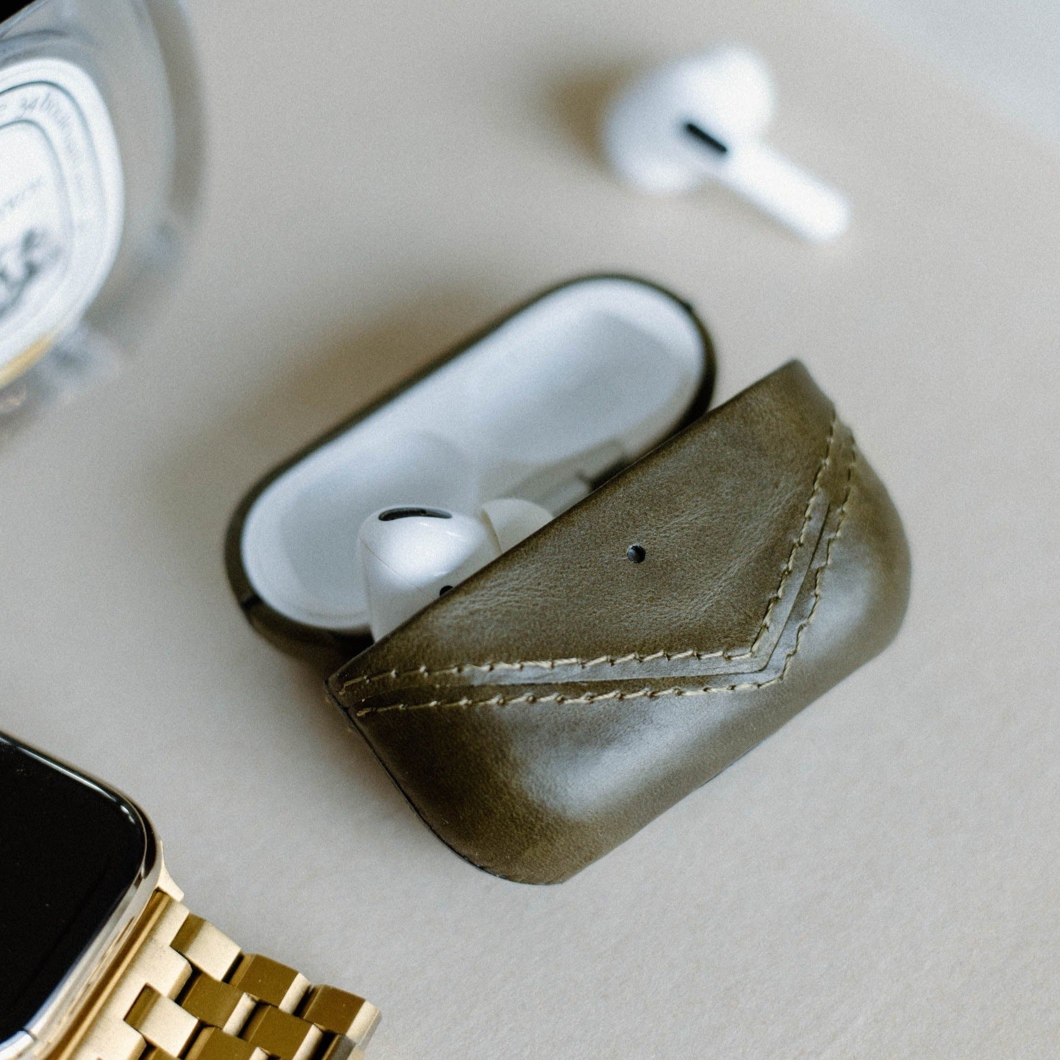Open leather AirPods case in olive green with AirPods inside, next to a watch and a candle.