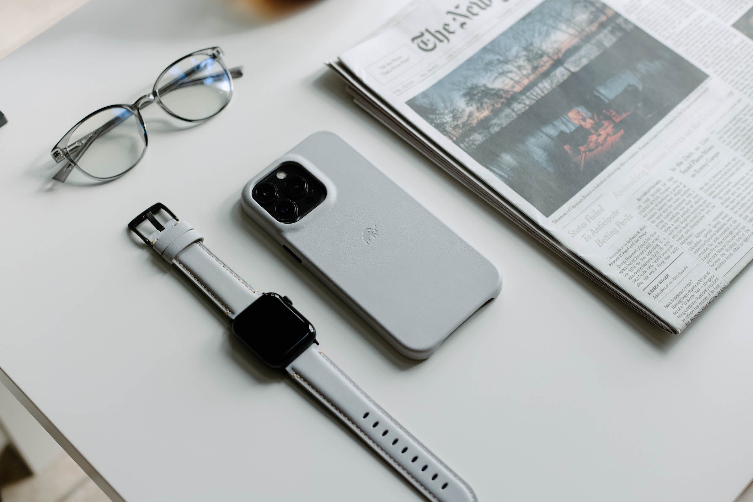 A light gray phone case next to a smartwatch and a newspaper on a table.