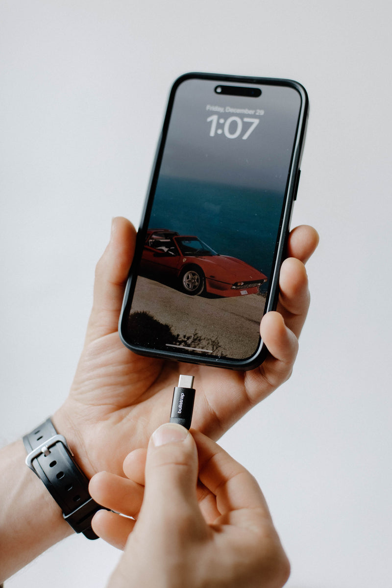 Hand holding phone with red sports-car wallpaper and lockscreen text 'Friday, December 29 1:07'; fingers holding a USB-C adapter below.