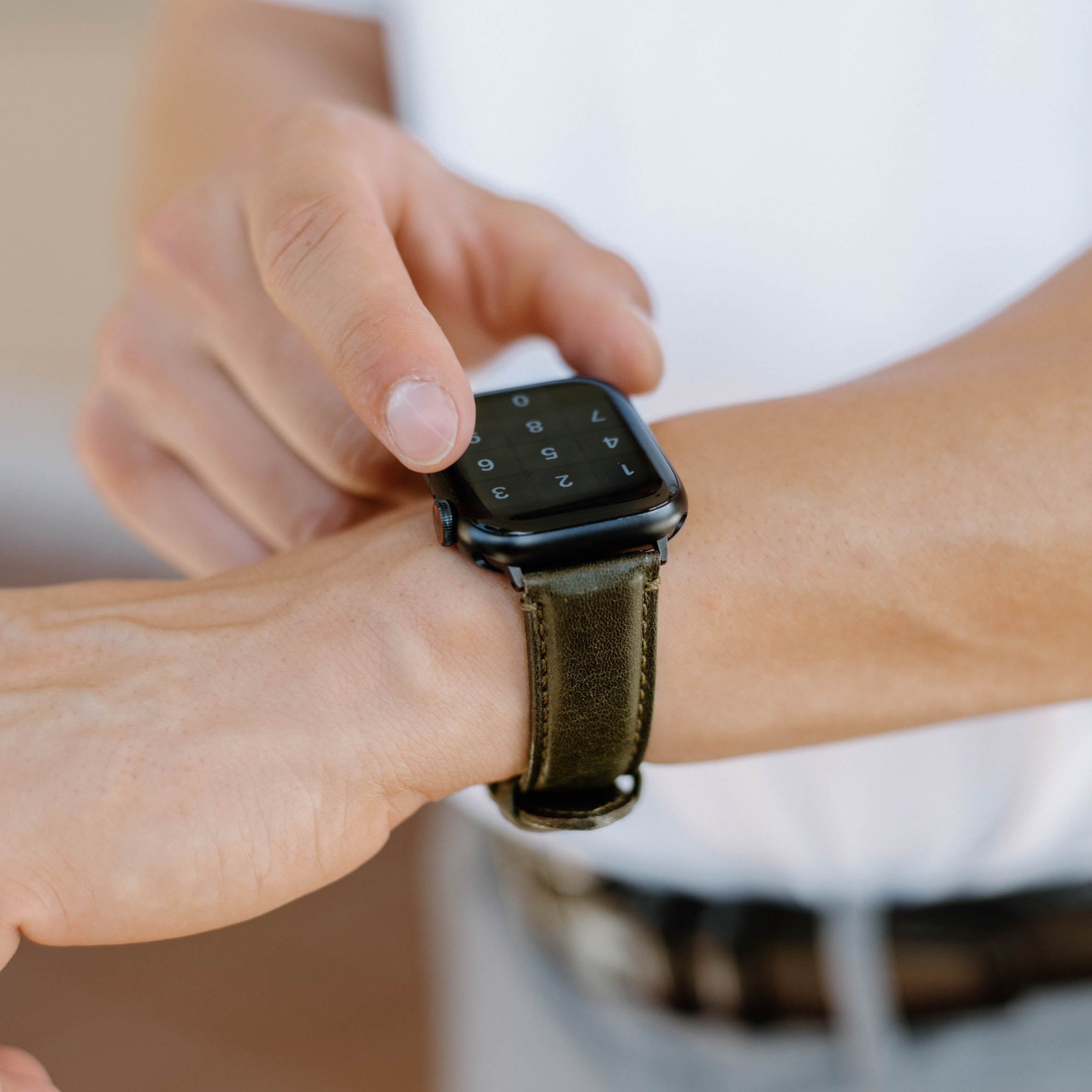 Person tapping Apple Watch with olive leather strap; watch screen shows keypad: 1 2 3 4 5 6 7 8 9 0