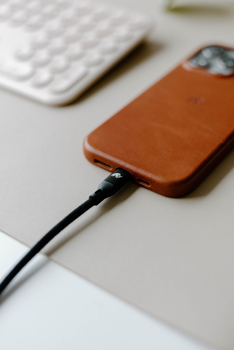 Brown leather-cased smartphone on a desk with a black braided charging cable plugged into its port.