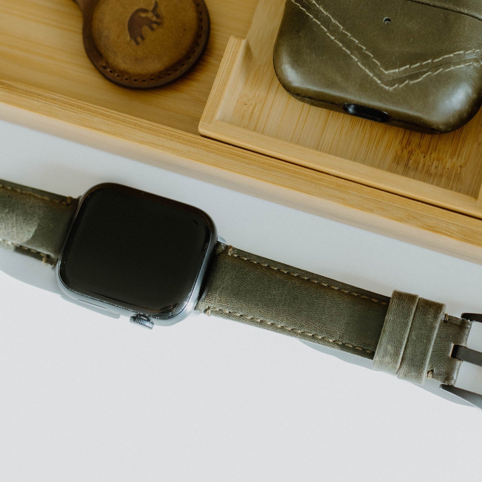 Olive leather Apple Watch with Maverick strap laid diagonally next to a wooden tray holding leather accessories