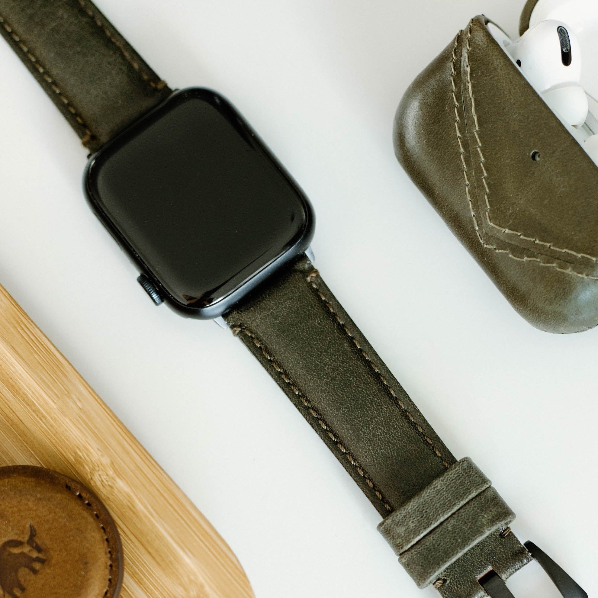 A black smartwatch with a green leather strap next to a green AirPods case and a wooden surface.