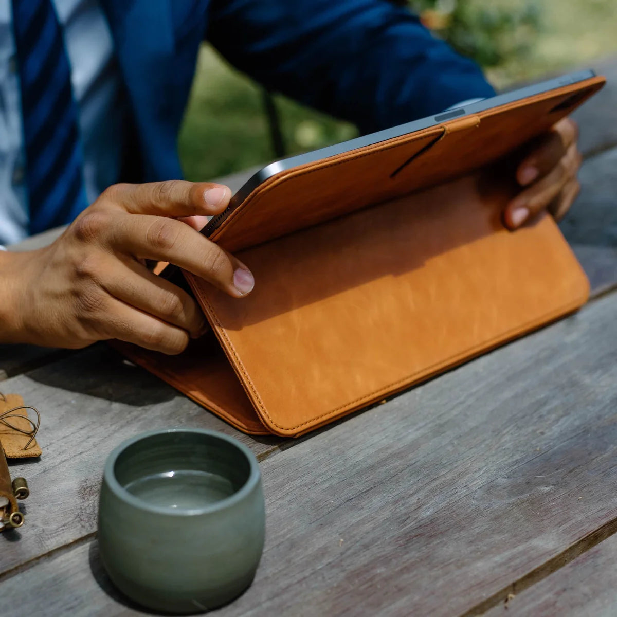 Hands holding tablet in tan leather folio on a wooden table, green ceramic cup in the foreground.