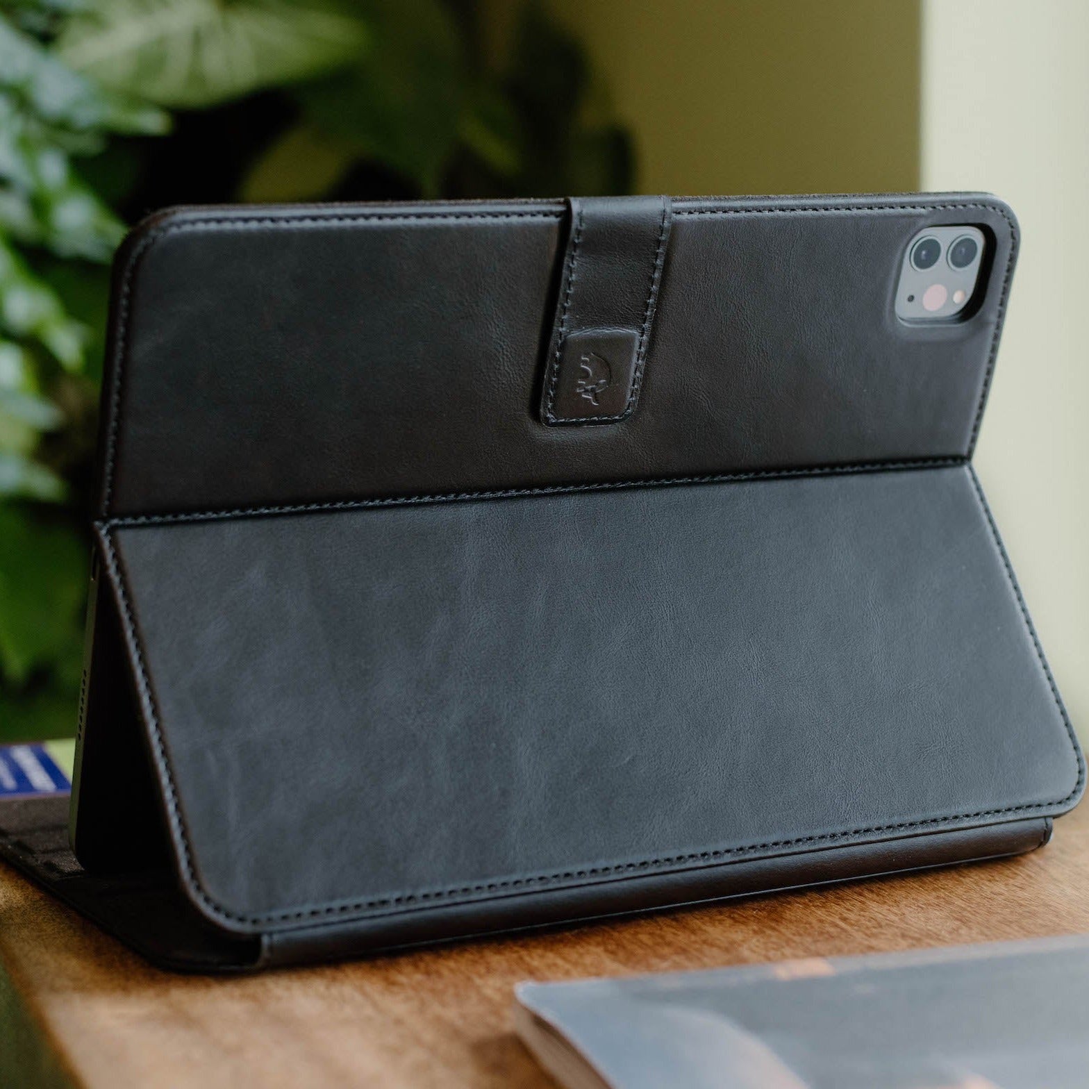 Black leather folio tablet case folded into a stand, camera cutout visible, resting on a wooden table.