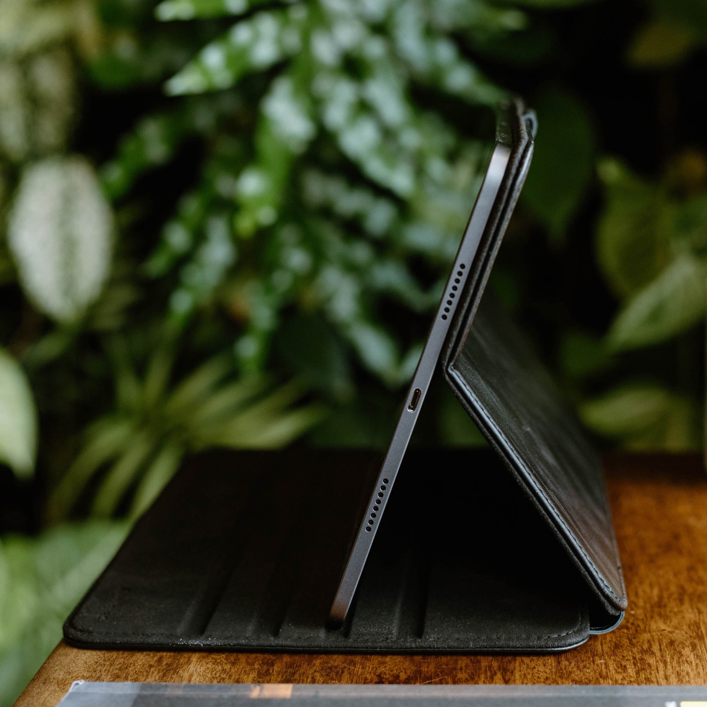 Side view of a black leather iPad case on a wooden surface with greenery in the background.