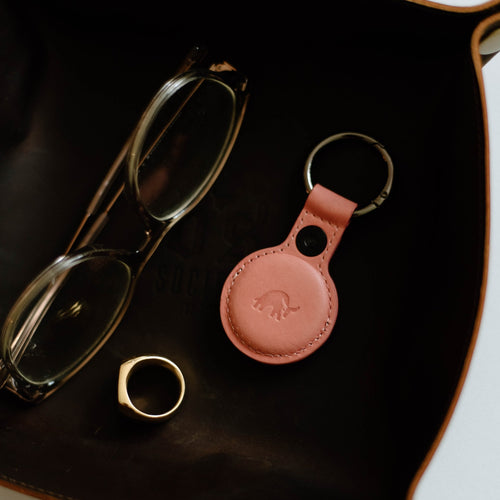 A pink leather AirTag holder on a tray with glasses and a ring.