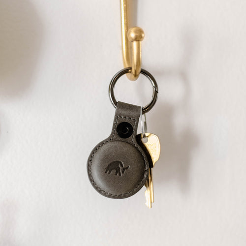 Slate gray leather BullTag AirTag key fob hanging from a brass hook with a small gold key attached