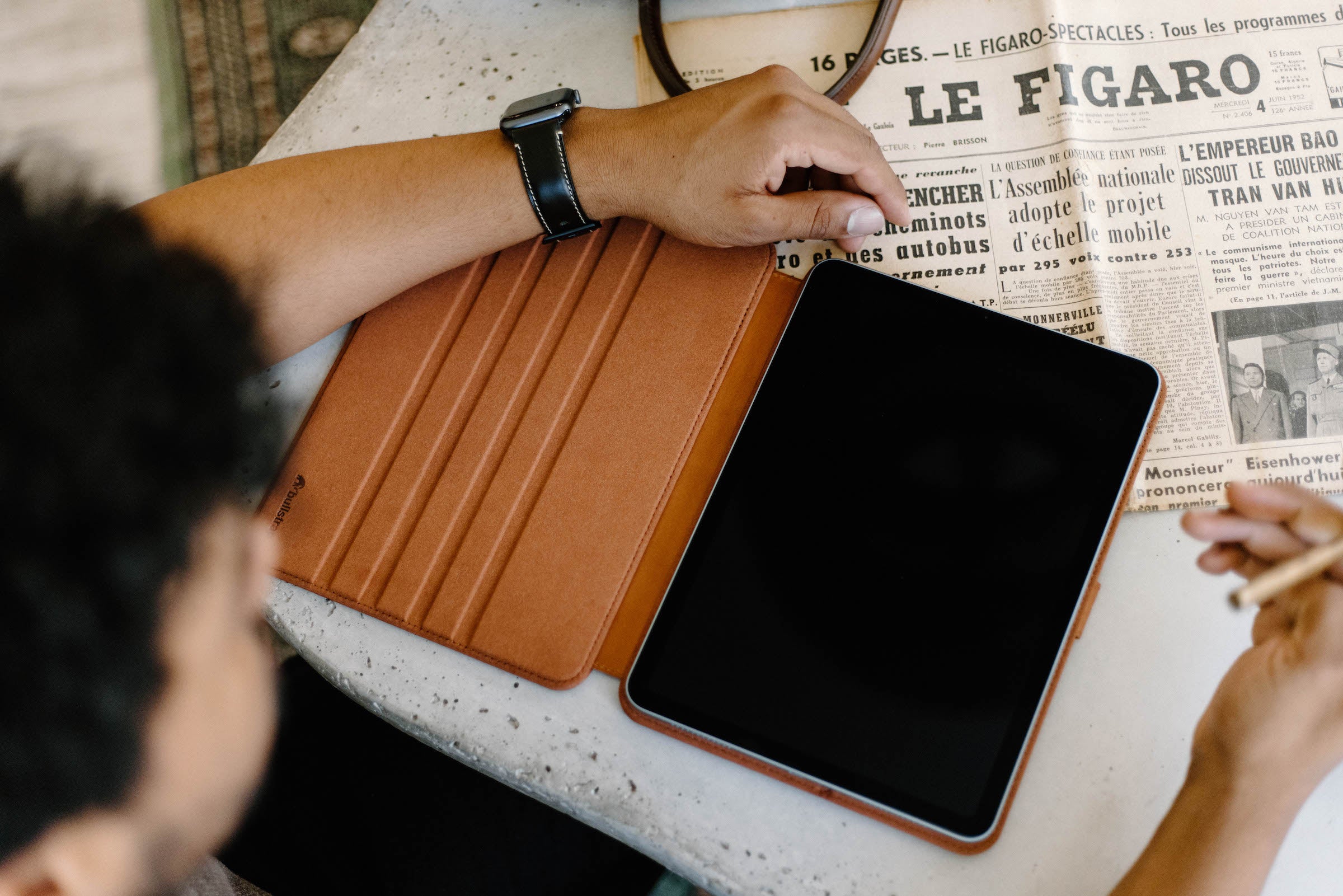Tablet in brown folio on a table beside a newspaper headlined 'LE FIGARO', two hands and a stylus visible.