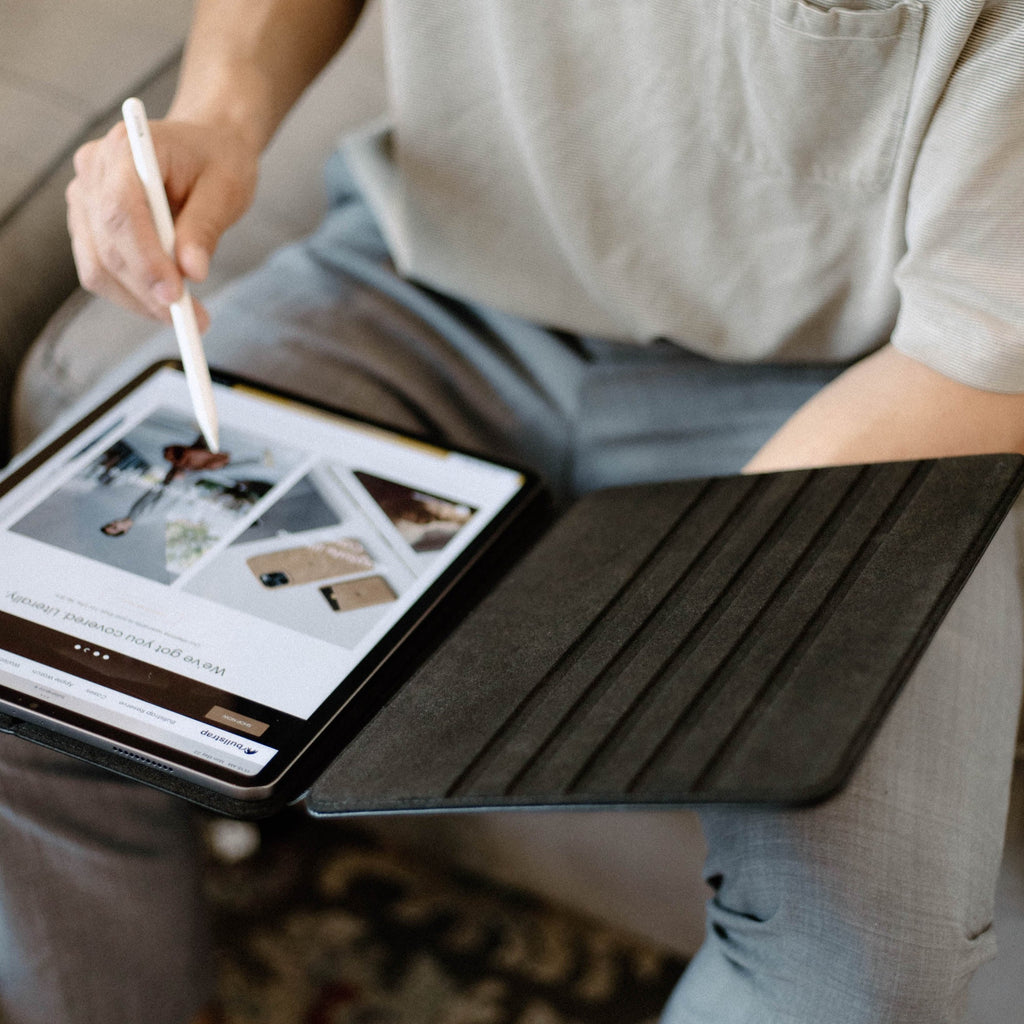 Black leather folio iPad case open on a person's lap with Apple Pencil touching the screen; screen text reads "We've got you covered".