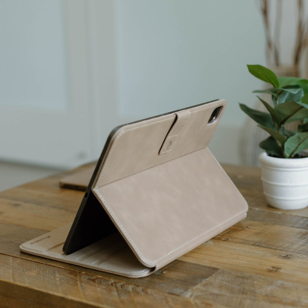 Leather iPad Case in Dune color, displayed on a wooden table with a plant in the background.