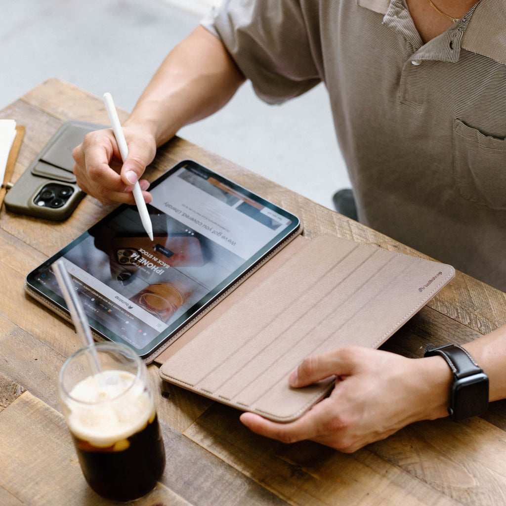 A person using a DUNE leather iPad case while writing on the iPad with a stylus, sitting at a wooden table with a drink.