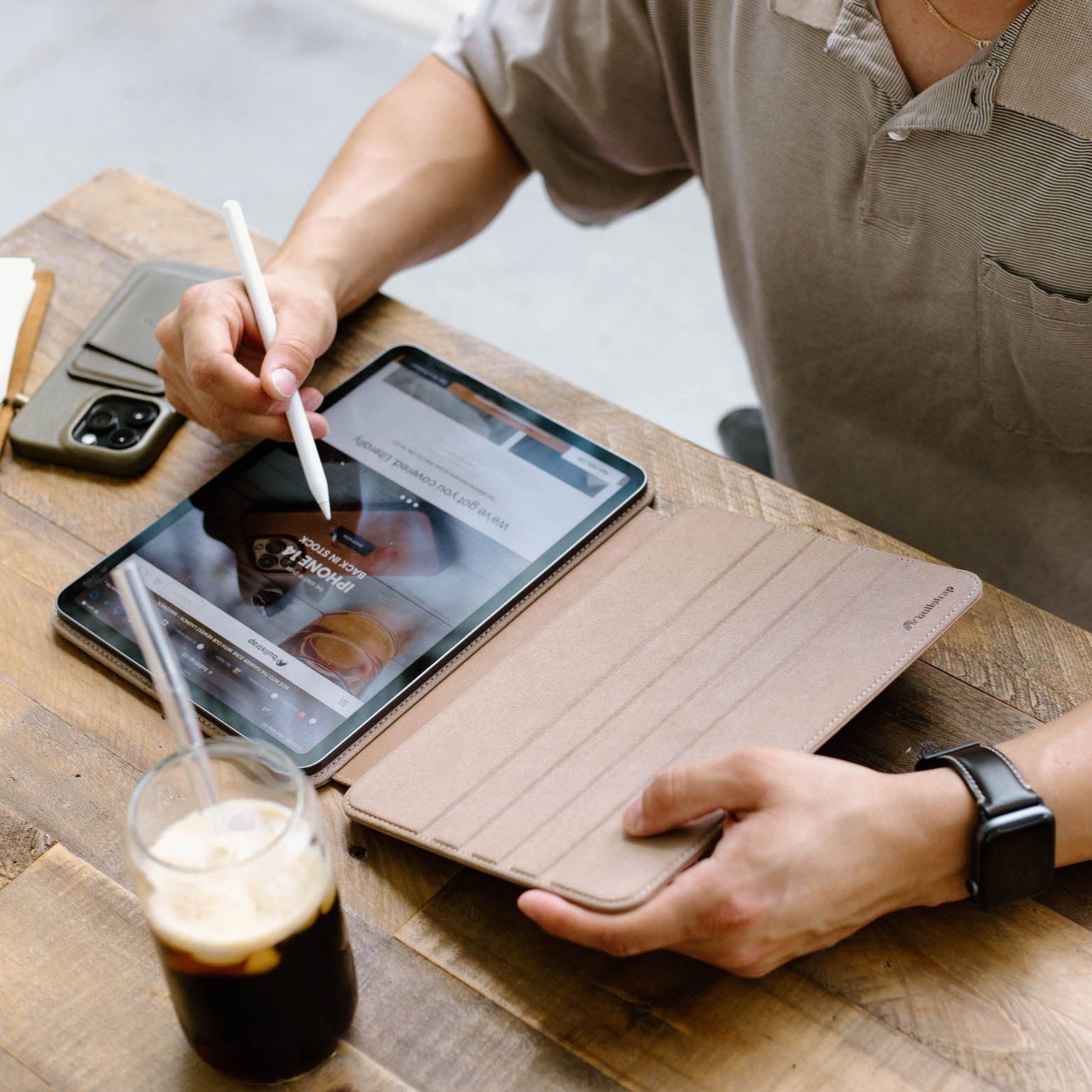 A person using a DUNE leather iPad case while writing on the iPad with a stylus, sitting at a wooden table with a drink.