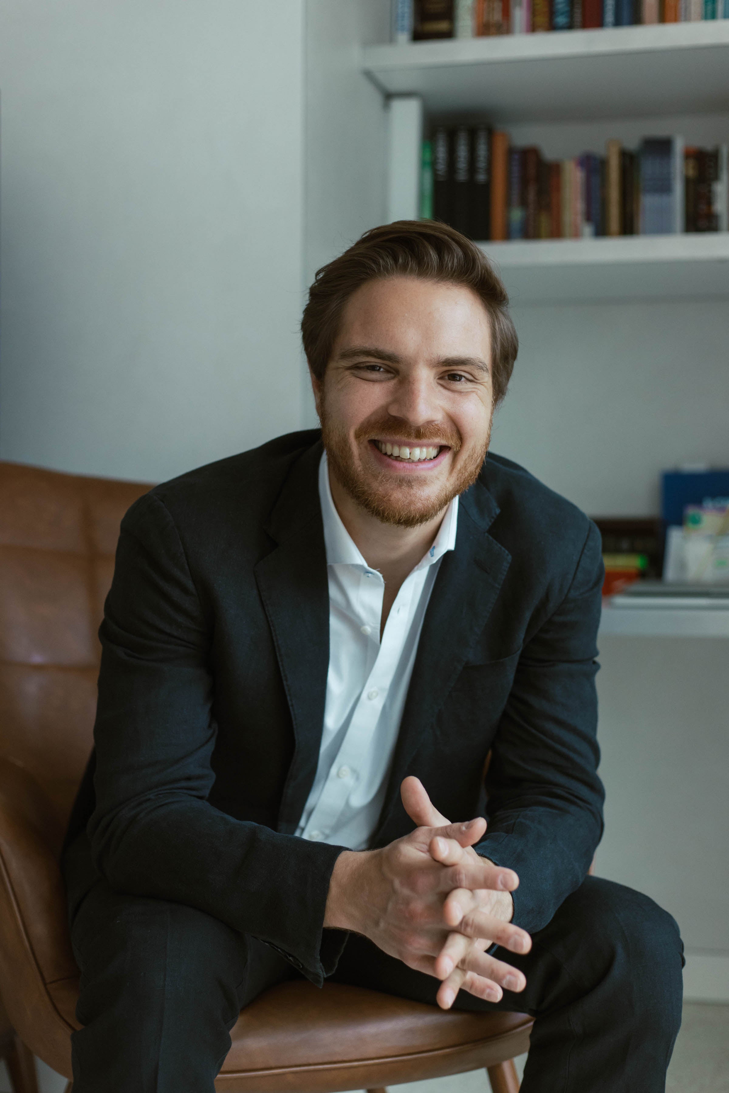 Young man in dark suit jacket and white shirt sitting on a leather chair, smiling and leaning forward with clasped hands.