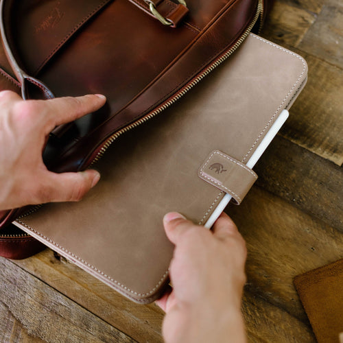 A person placing a Leather iPad Case - DUNE into a brown leather bag.